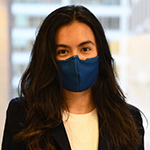 Female (Jacqueline Enriquez) with dark long hair wearing a dark-colored blazer, light-colored shirt and a dark mask covering her nose and mouth.